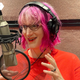 Author Charlie Jane Anders is seen recording the audiobook for her latest release, Never Say You Can't Survive. She's wearing a bright orange t-shirt and has neon pink hair.